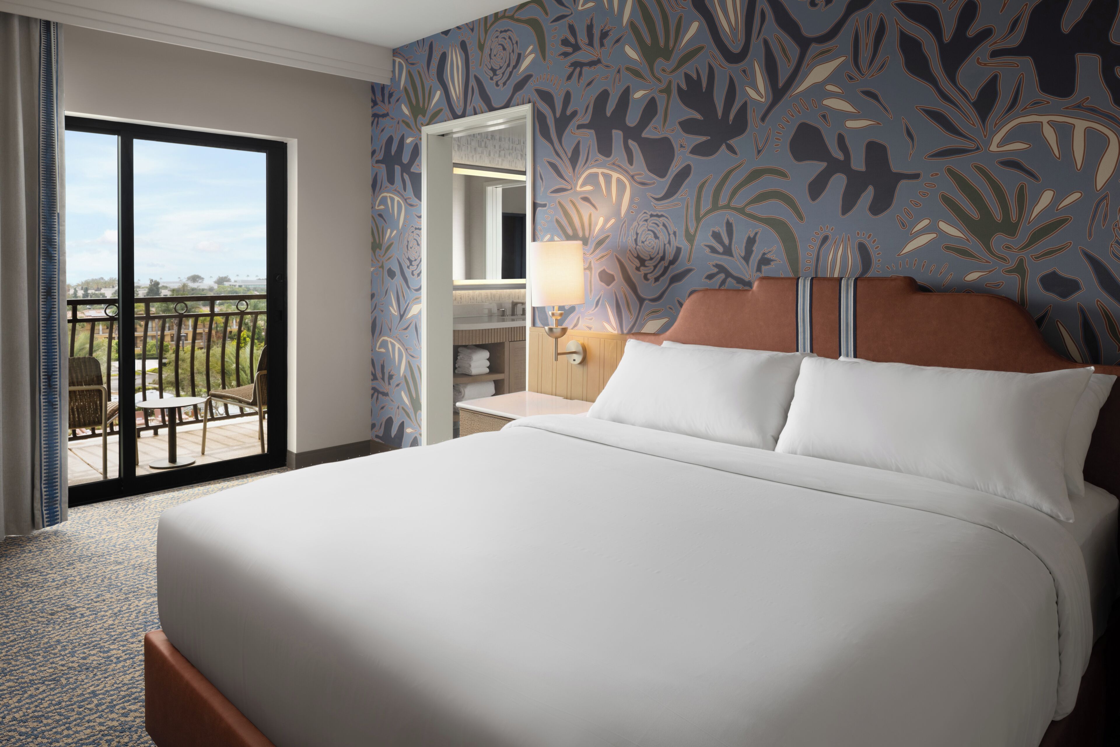 A hotel room with a large bed, a bathroom with a mirror, and a balcony with outdoor furniture.