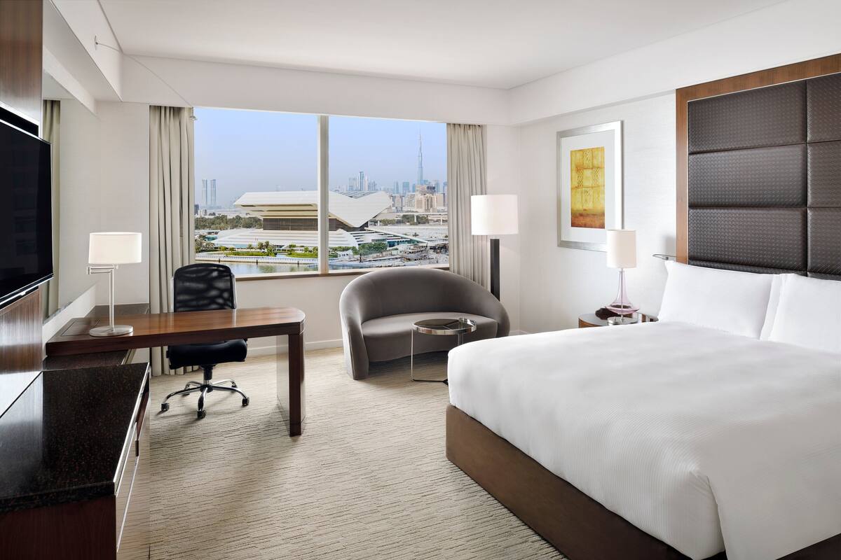 A hotel room with a large bed, a desk, a chair, a TV, and a view of a cityscape.