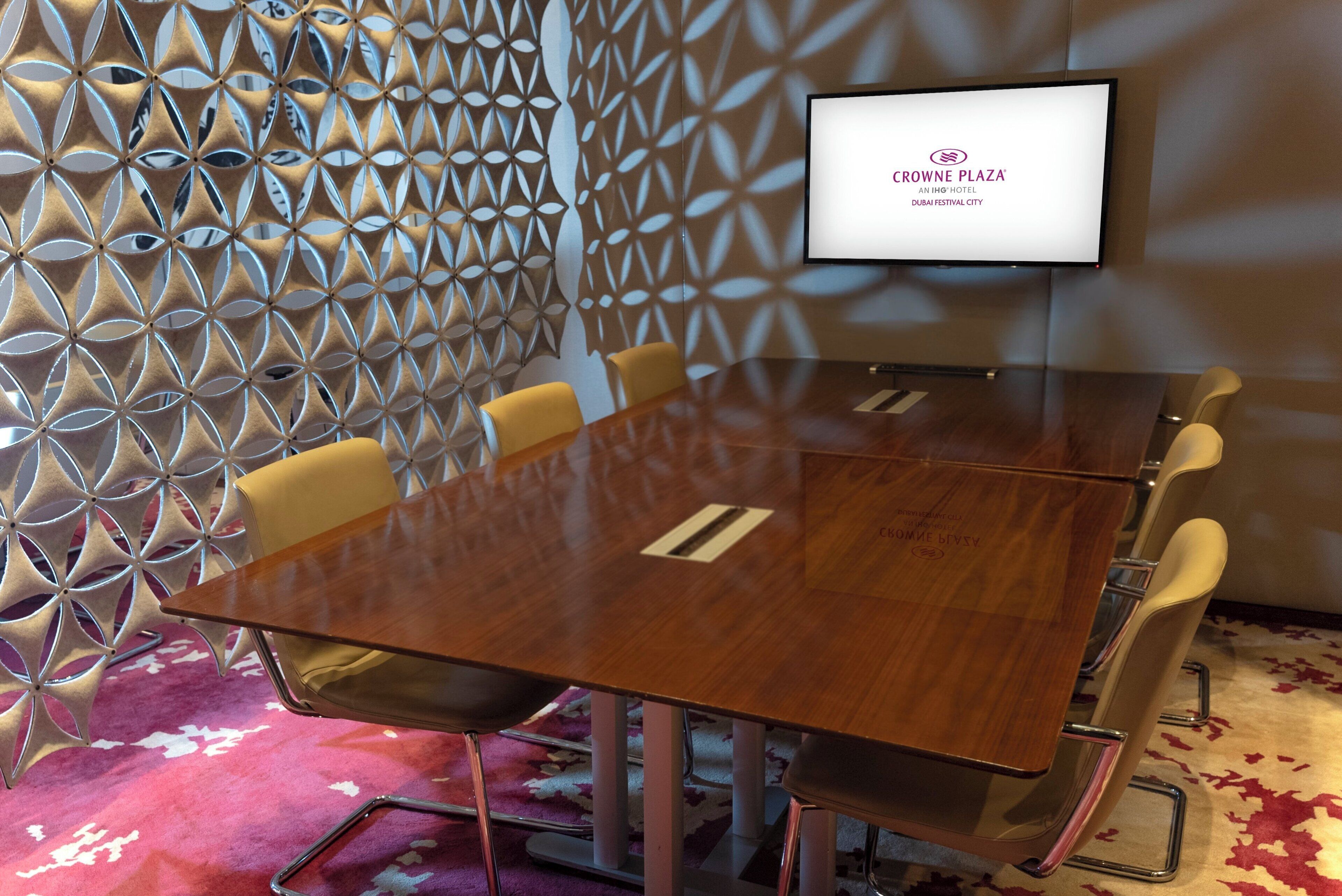 Une salle de conférence avec une table en verre, quatre chaises et un panneau décoratif au mur. Un écran affiche le logo de Crowne Plaza.