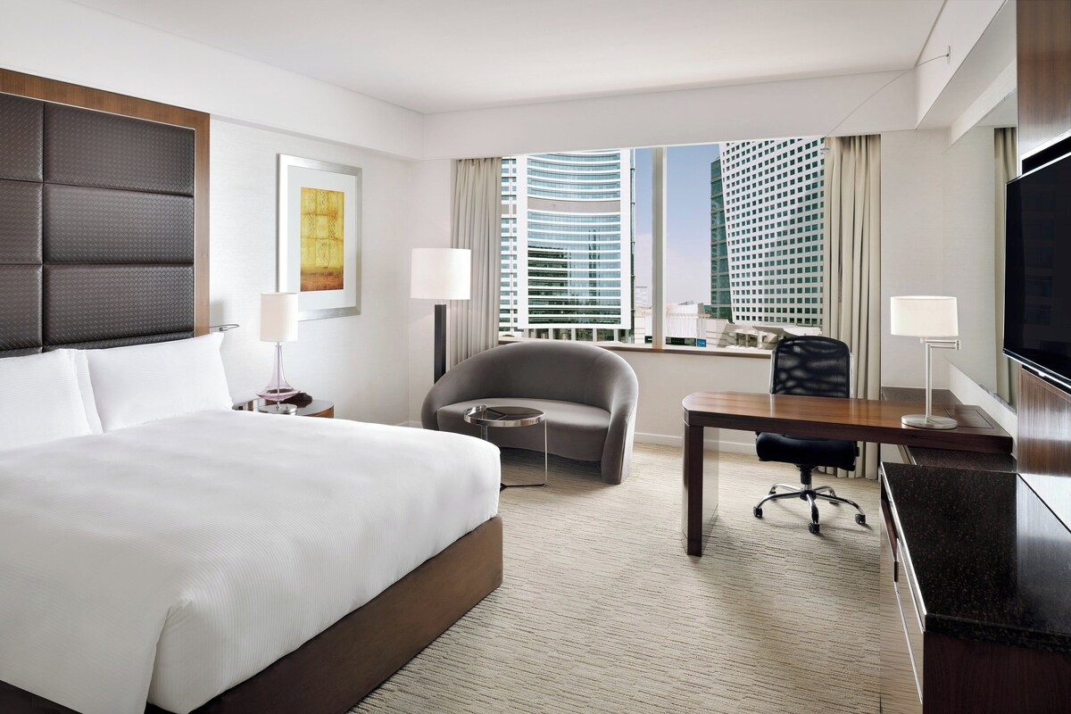 Ein Hotelzimmer mit einem großen Bett, einem Schreibtisch, einem Sessel und einem Fernseher, mit Blick auf hohe Gebäude.