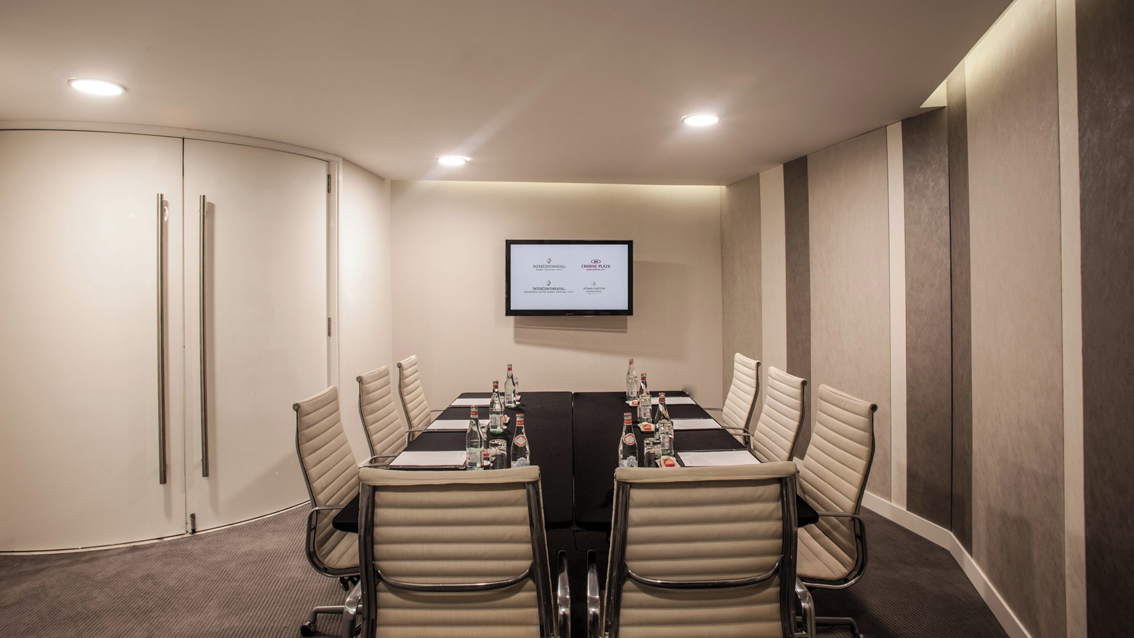 A conference room with a long table, six chairs, a flat-screen TV on the wall, and a closed door.