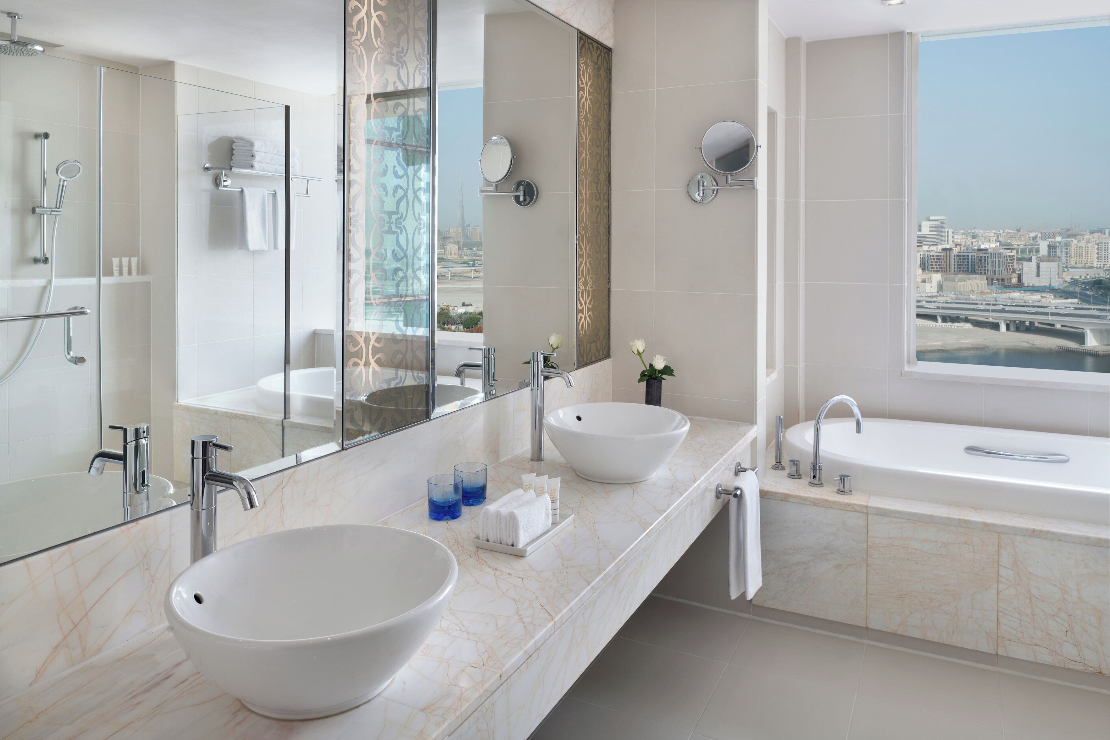 A modern bathroom with a large mirror, two sinks, a bathtub, and a city view.