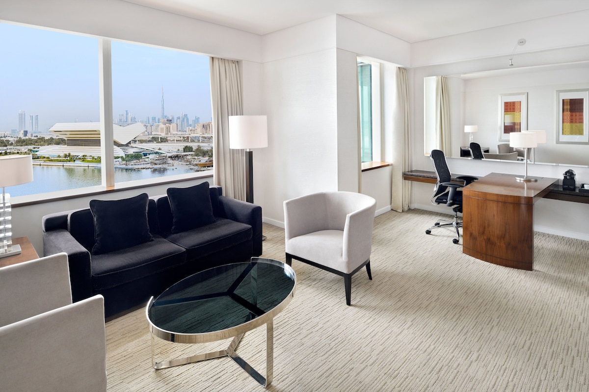 Een moderne hotelkamer met een groot raam met uitzicht op de stad, een zwarte bank, een witte fauteuil, een glazen salontafel en een bureau met stoel.