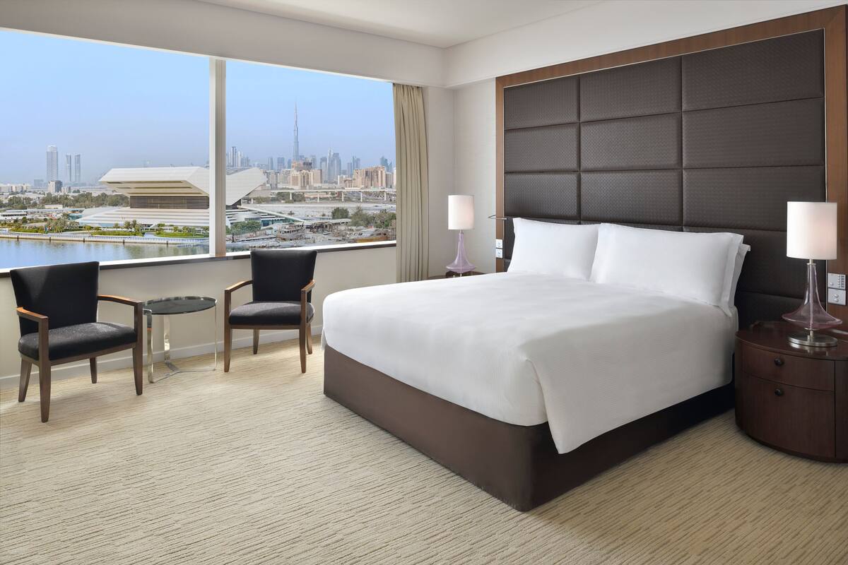A hotel room with a large bed, two chairs, a small table, and a view of a cityscape.