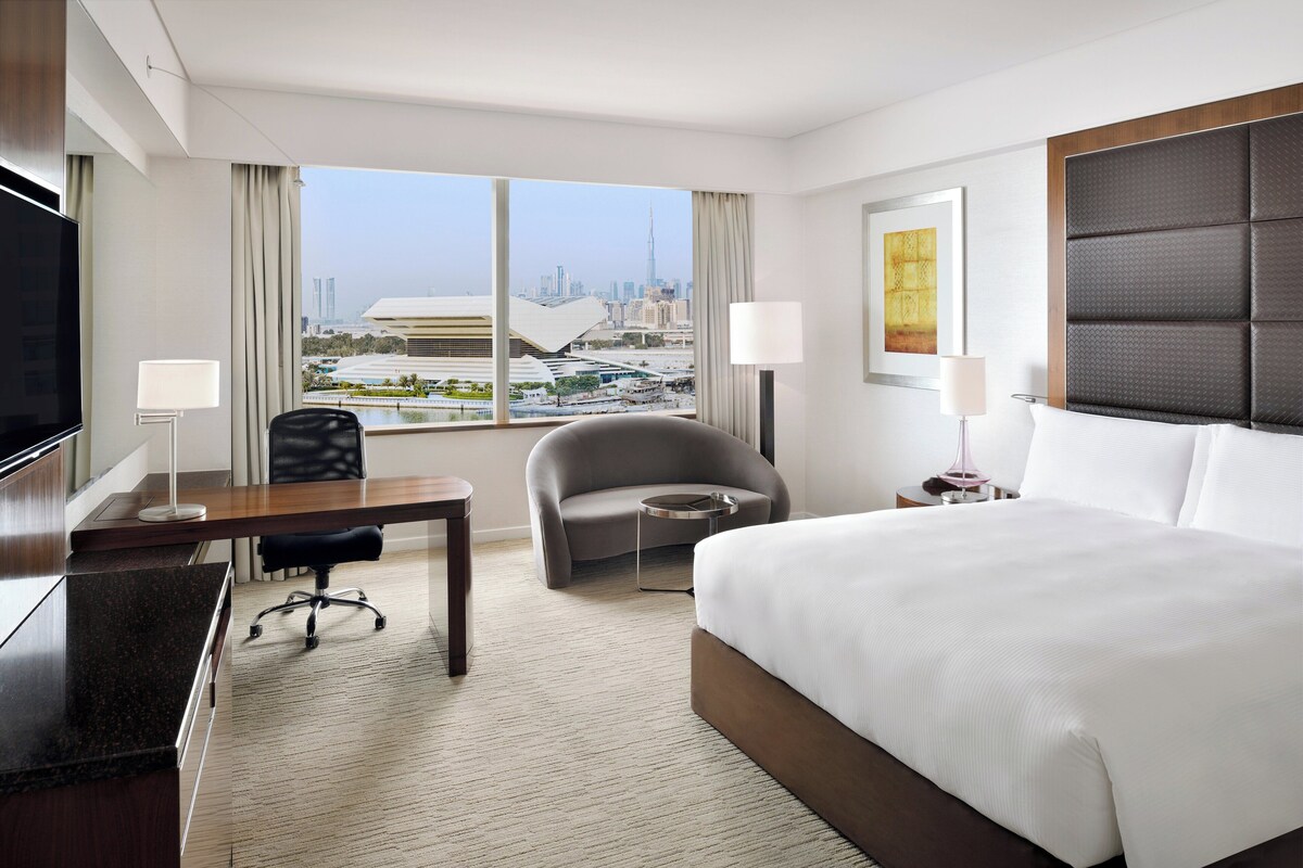 Ein Hotelzimmer mit einem großen Bett, einem Schreibtisch, einem Sessel, einem Fernseher und Blick auf eine Stadtlandschaft.