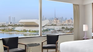 Habitación de hotel con un ventanal grande que ofrece vistas del horizonte urbano, incluyendo un rascacielos destacado. Hay dos sillas y una mesita en la habitación.