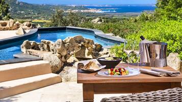 A poolside dining area with a view of the sea, a wooden table with a fruit plate, a bowl, and a bottle of champagne.