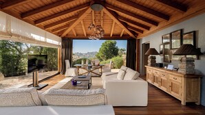 Un vaste espace de vie doté d’un plafond en bois, d’une grande fenêtre offrant une vue sur l’extérieur, d’un ensemble de canapés et d’un meuble en bois agrémenté d’objets décoratifs.