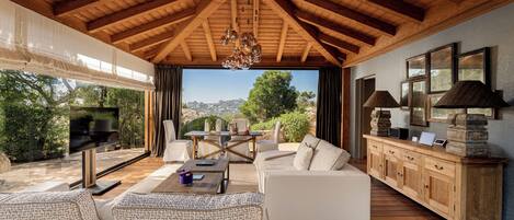 A spacious living area with a wooden ceiling, a large window offering a view of the outdoors, a sofa set, and a wooden cabinet with decorative items.