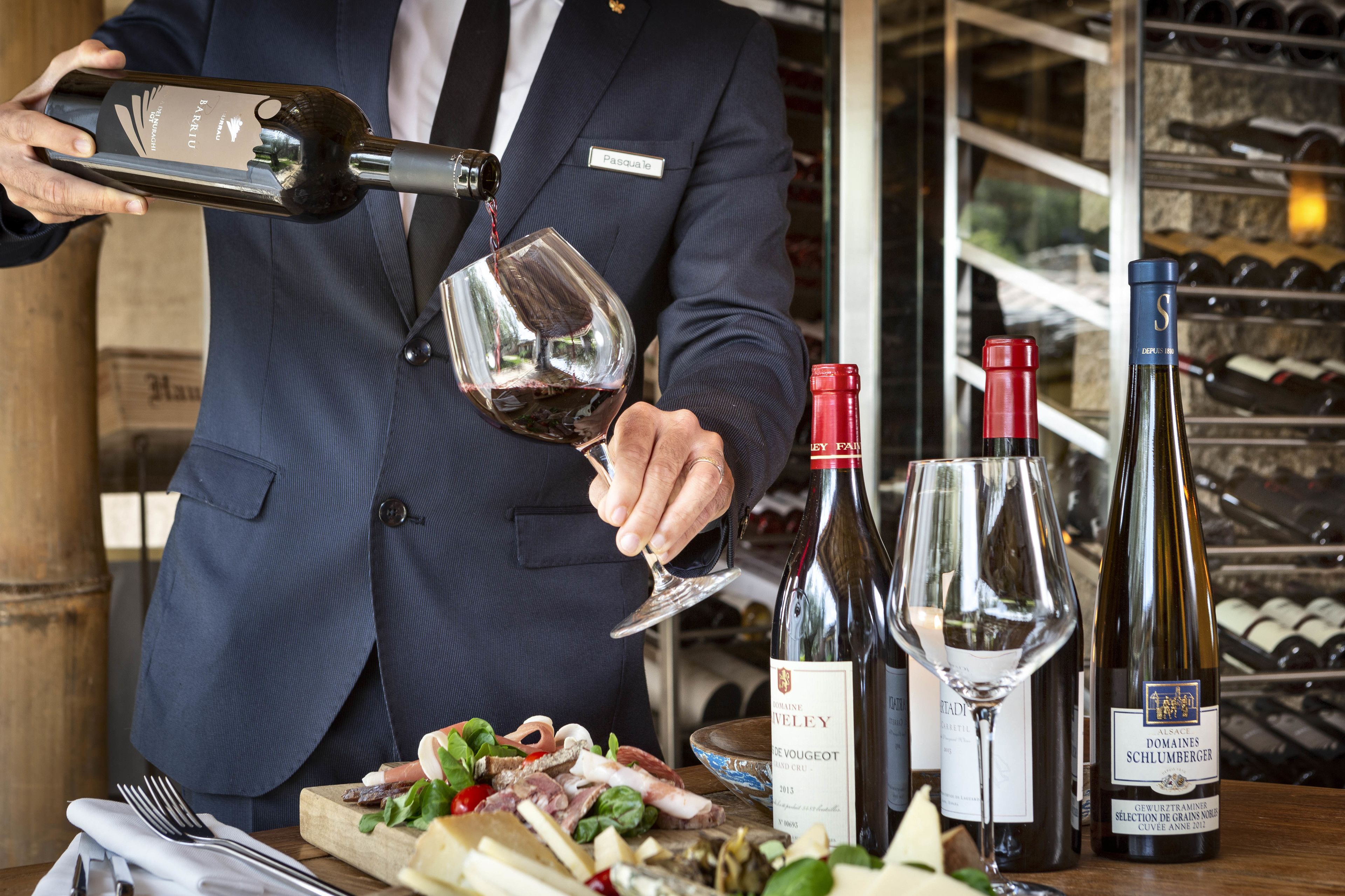 Eine Person gießt Wein aus einer Flasche ins Glas. Im Hintergrund sind Weinflaschen und ein Weinkühler zu sehen.