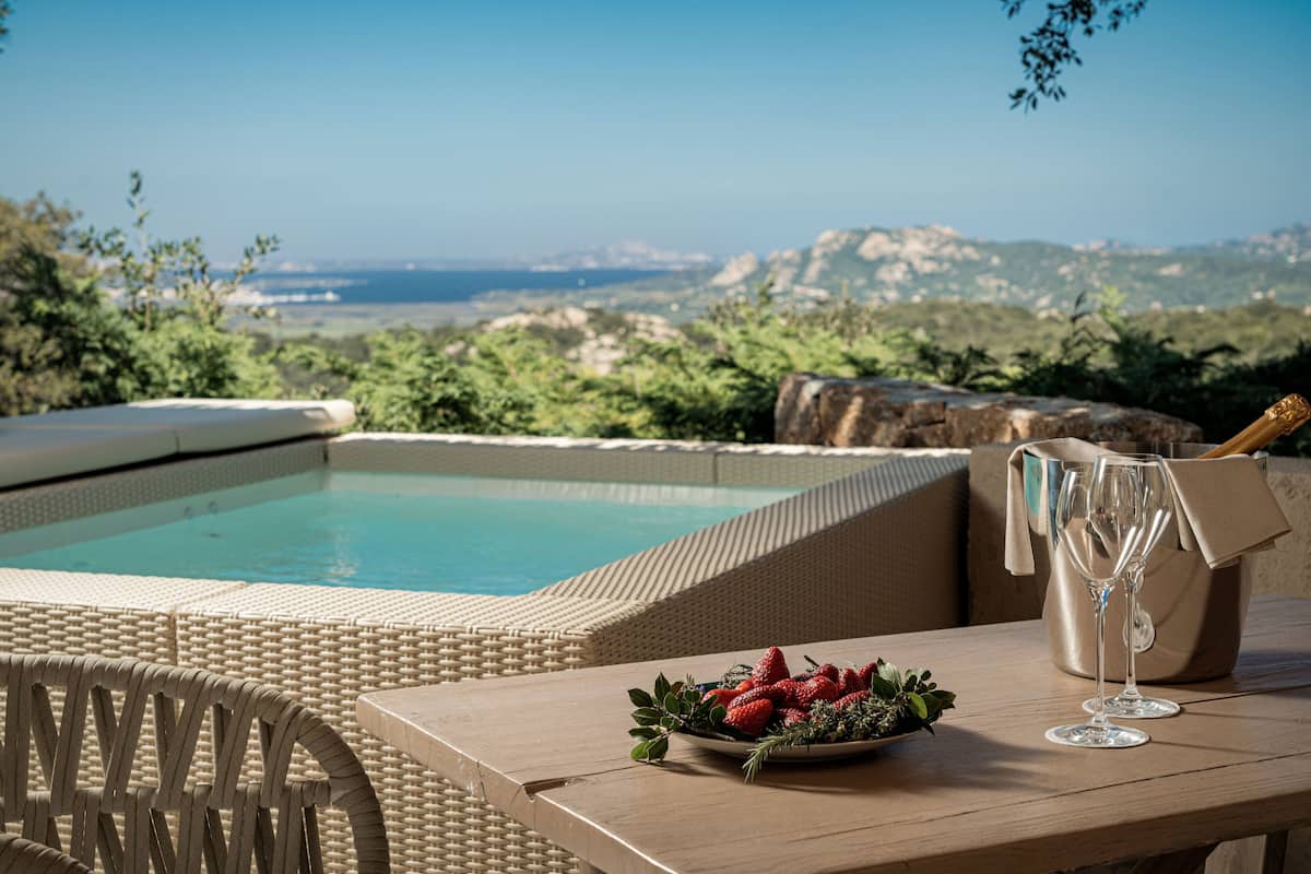 A poolside setting with a wicker lounge chair, a table with a bowl of strawberries and a glass of champagne, and a view of the ocean and hills.