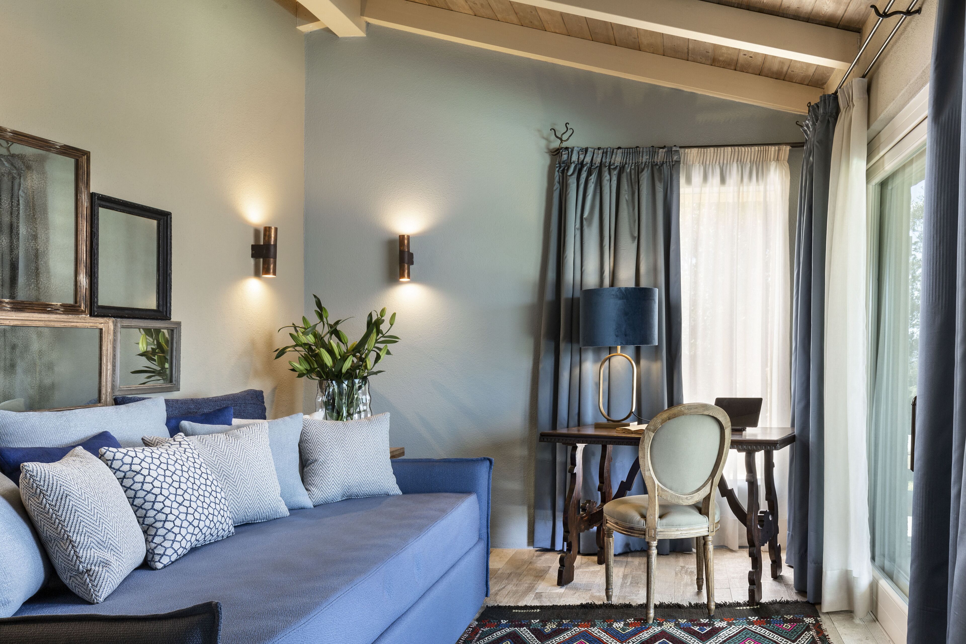Una sala acogedora con un sofá azul, una mesita con una lámpara y una silla. Hay luces en la pared y una planta sobre el sofá.