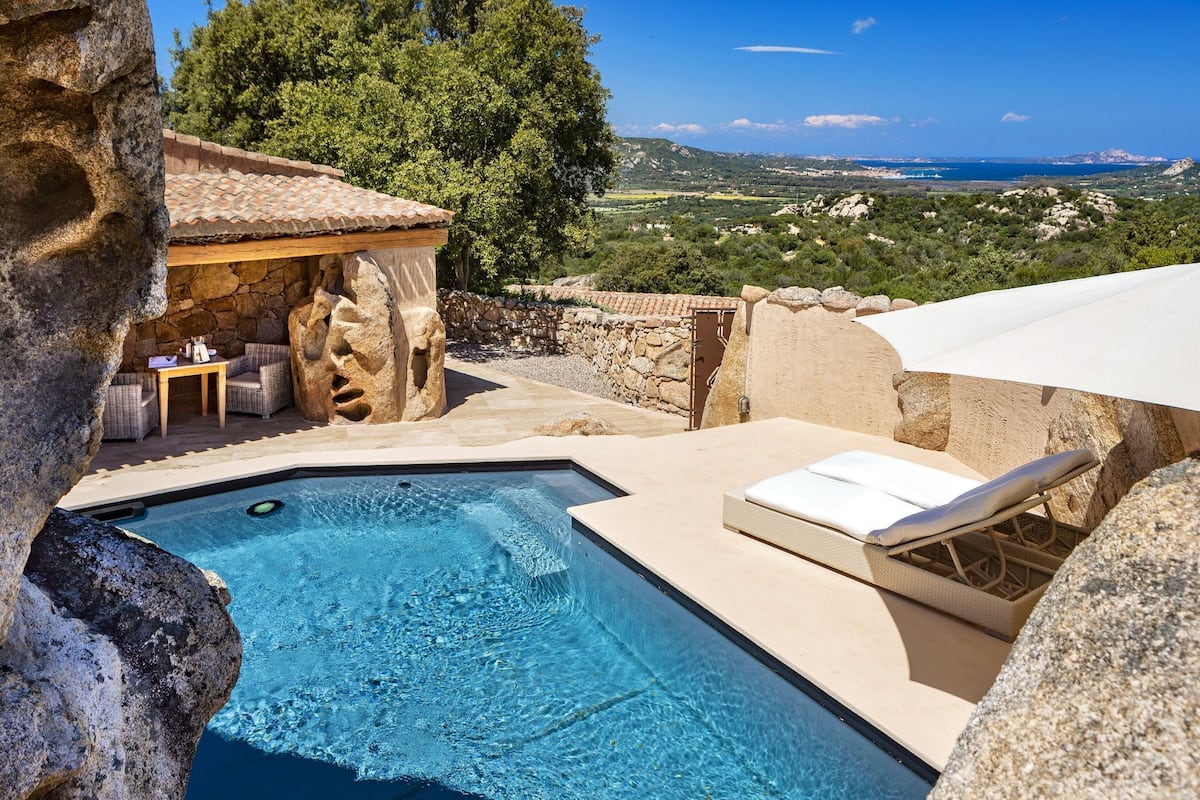 A pool area with a stone wall, a lounge chair, and a table with outdoor seating.