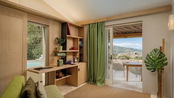 A modern living room with a wooden desk, a green sofa, a dining area with a table and chairs, and a view of the outdoors.