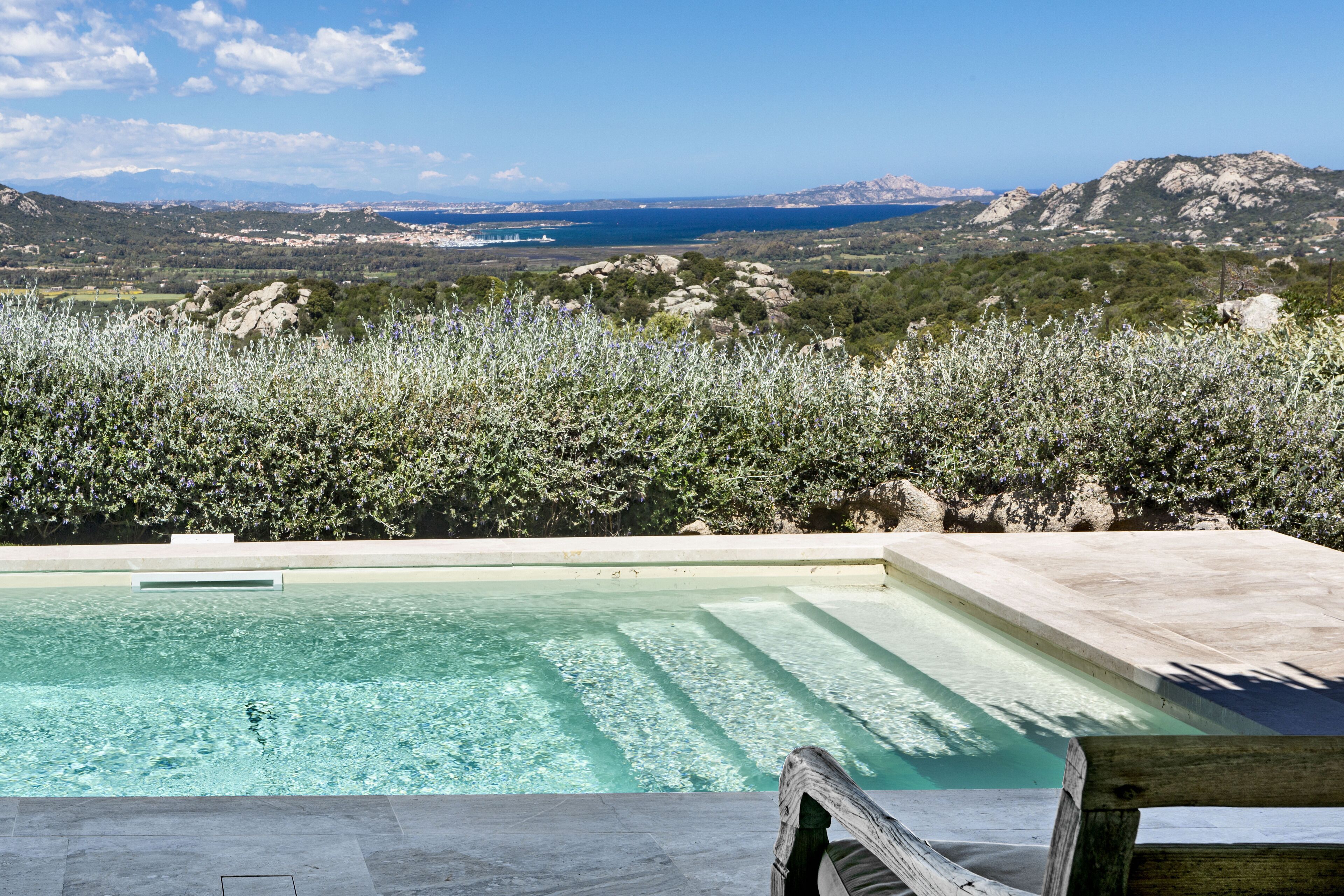 Ein Pool mit klarem Wasser, umgeben von üppiger Vegetation mit Blick auf eine malerische Landschaft aus Bergen und einem Gewässer.