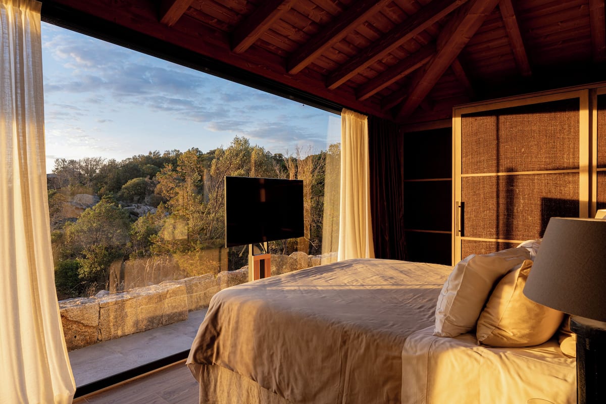 A bedroom with a bed, a television, and a view of a forested area.