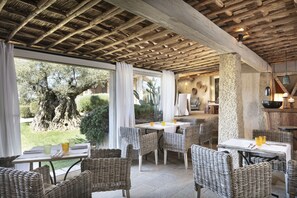 An outdoor dining area with wicker chairs and tables, a stone pillar, and a view of the garden. - Petra Segreta Resort & Spa (Olbia)