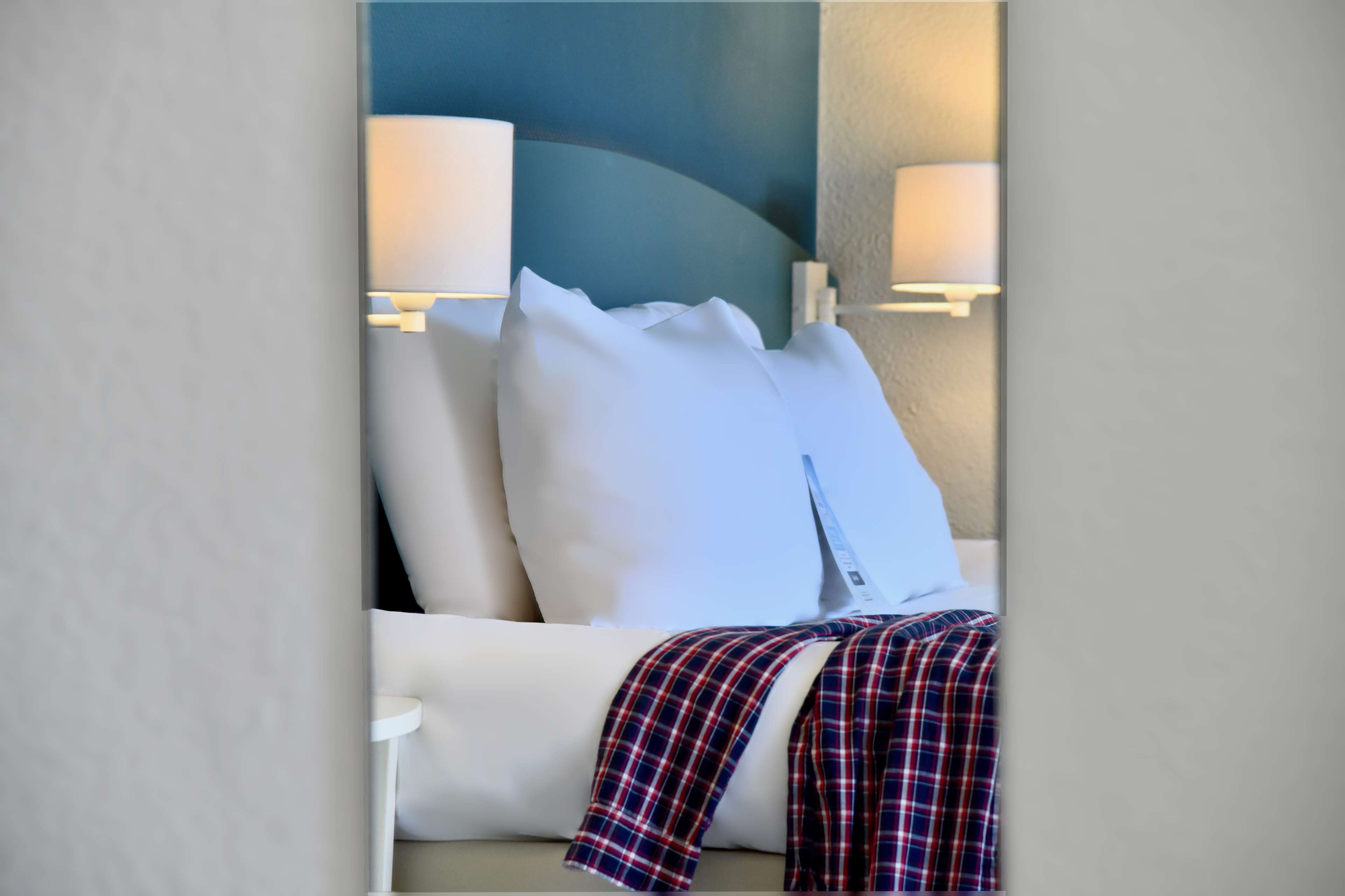A neatly made bed with white linens and a plaid blanket, two bedside lamps, and a wall-mounted light fixture.