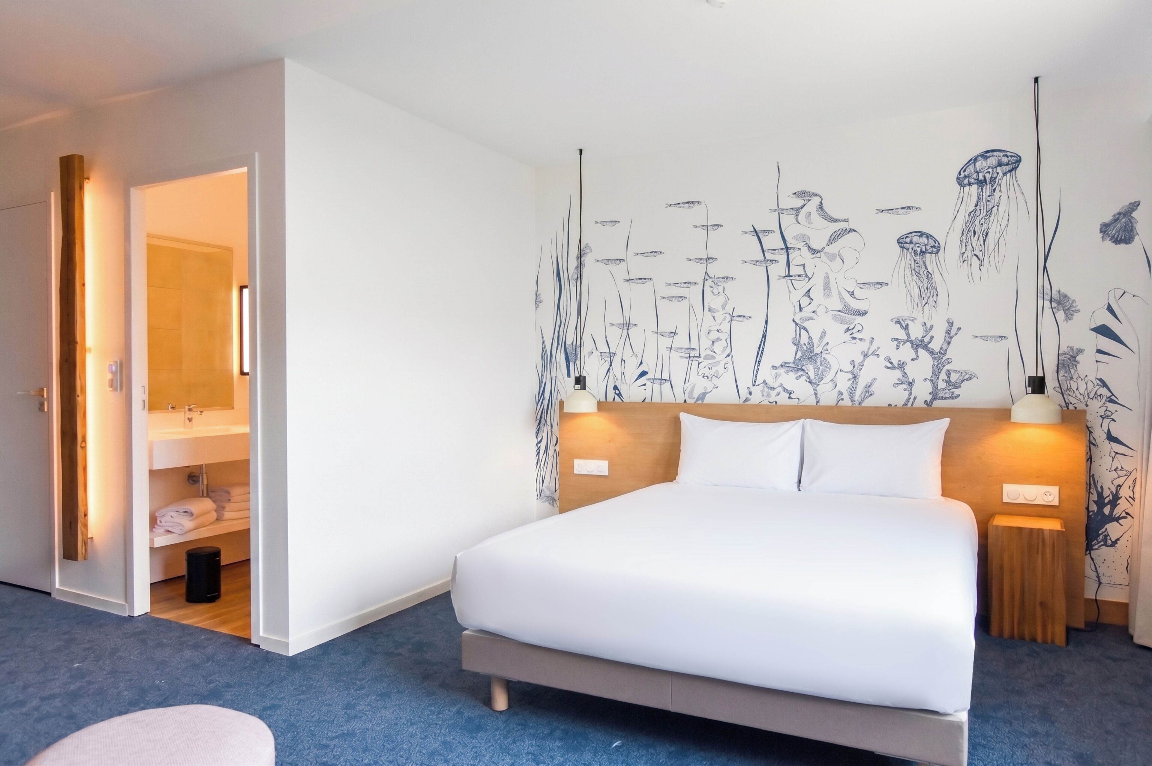 Une chambre d’hôtel avec un lit, une table de chevet, une lampe fixée au mur et une salle de bain visible à travers une porte ouverte.