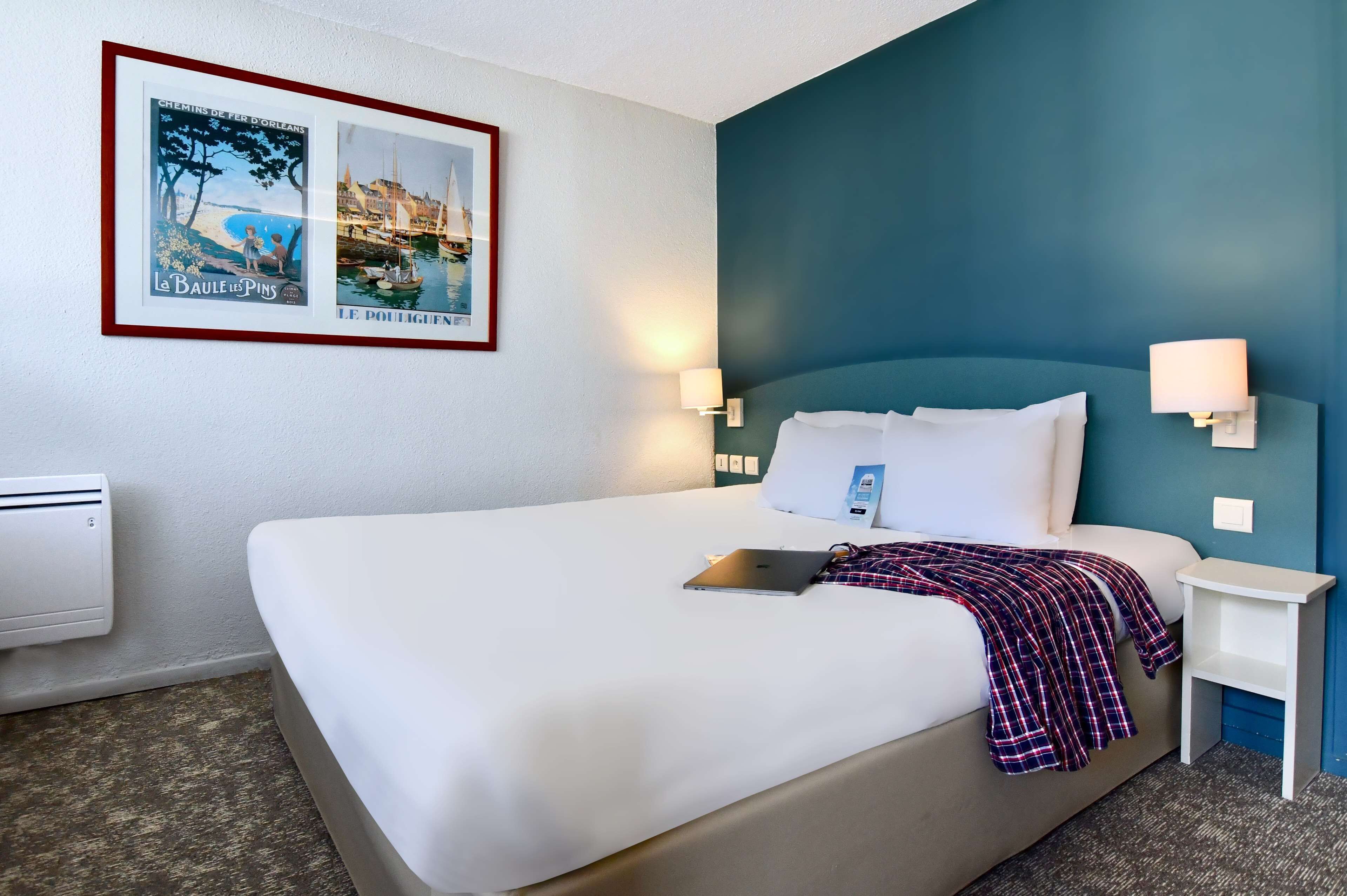 A hotel room with a large bed, two bedside tables, a wall-mounted air conditioner, and two framed pictures.