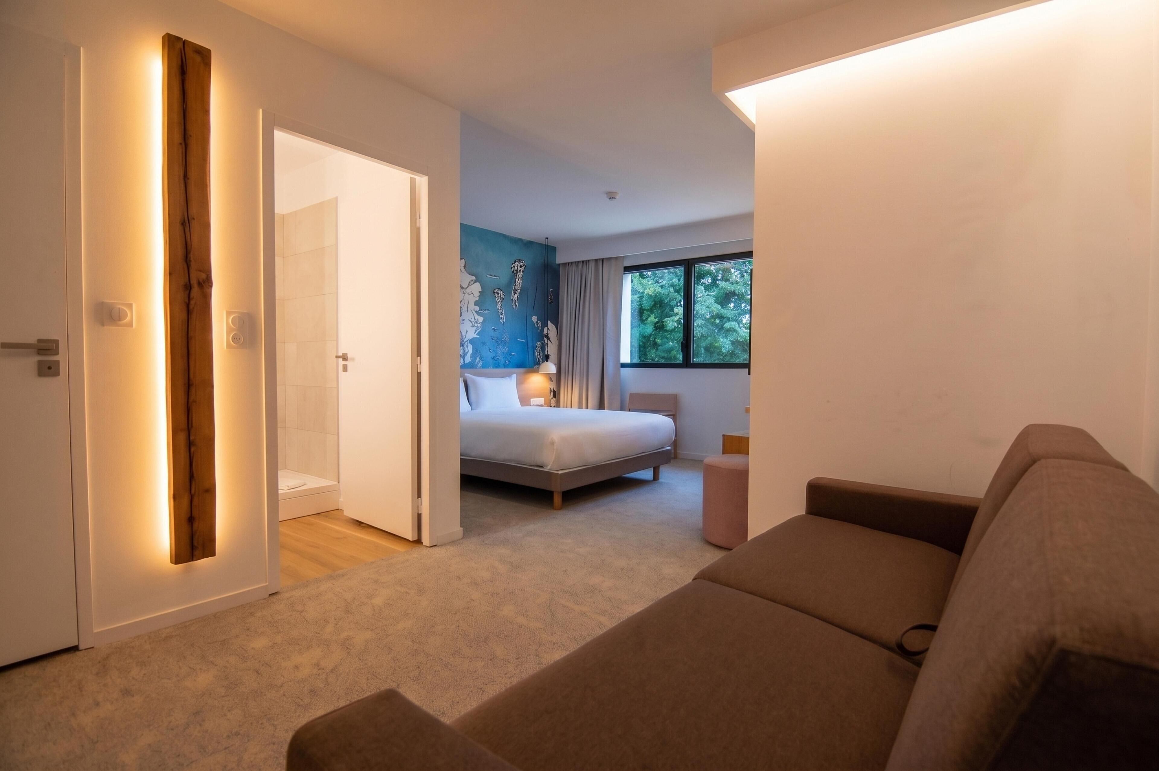 A modern hotel room with a large bed, a seating area, and a view of trees through the window.