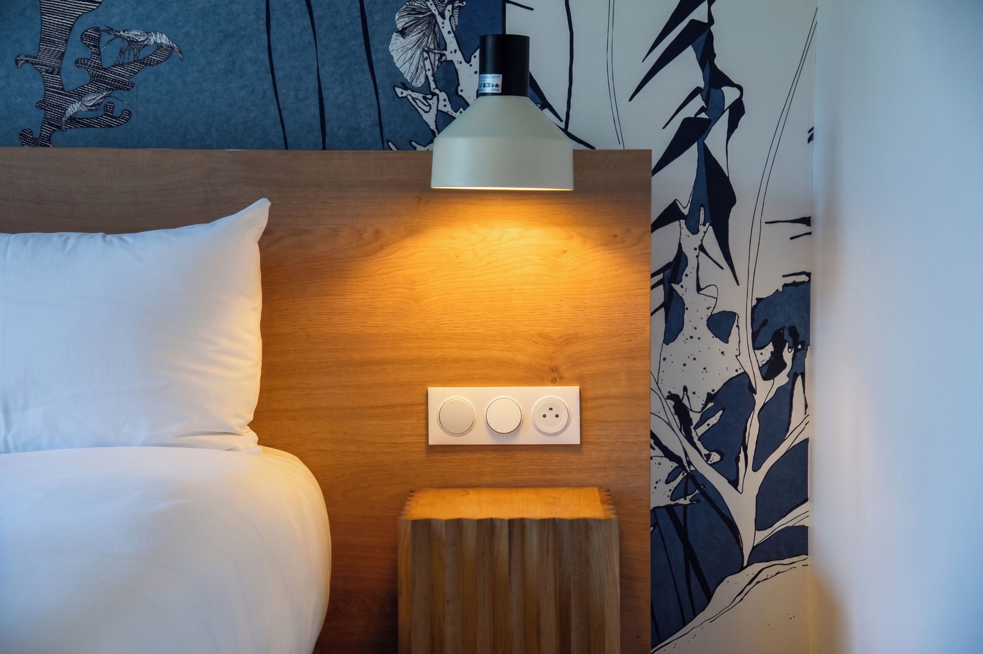 A hotel room with a bed, a bedside table with a lamp, and a power outlet.
