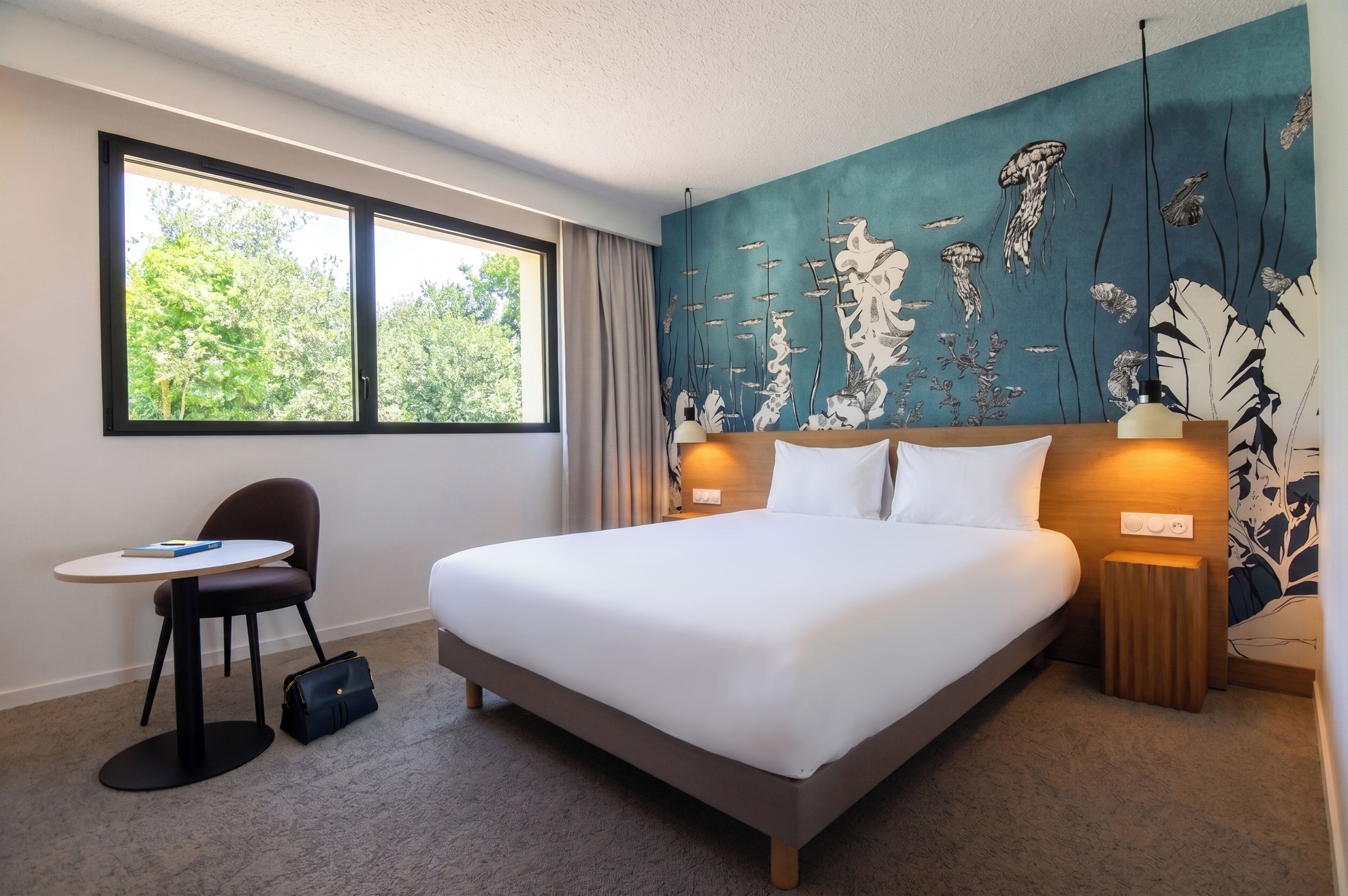 A hotel room with a bed, a desk, a chair, a handbag, and a window with a view of trees.