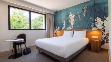A hotel room with a bed, a desk, a chair, a handbag, and a window with a view of trees.