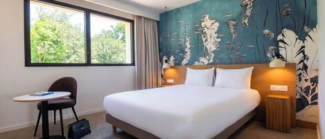 Habitación de hotel con cama, escritorio, silla, bolso y ventana con vista a árboles.