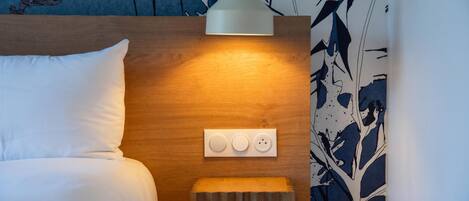 A hotel room with a bed, a bedside table, a lamp, and a power outlet.