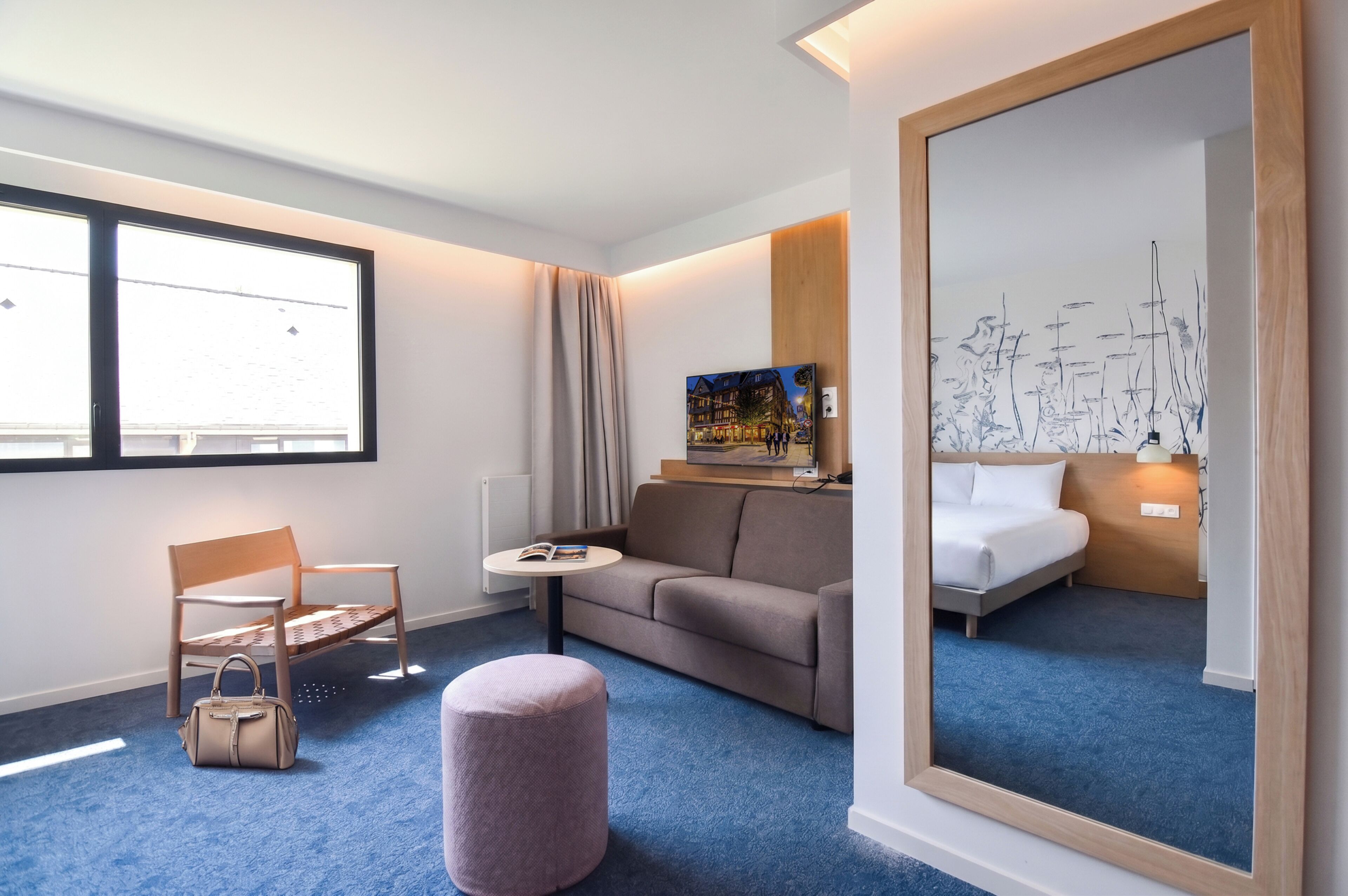 A hotel room with a bed, a sofa, a chair, a small table, a handbag, a mirror, and a window.