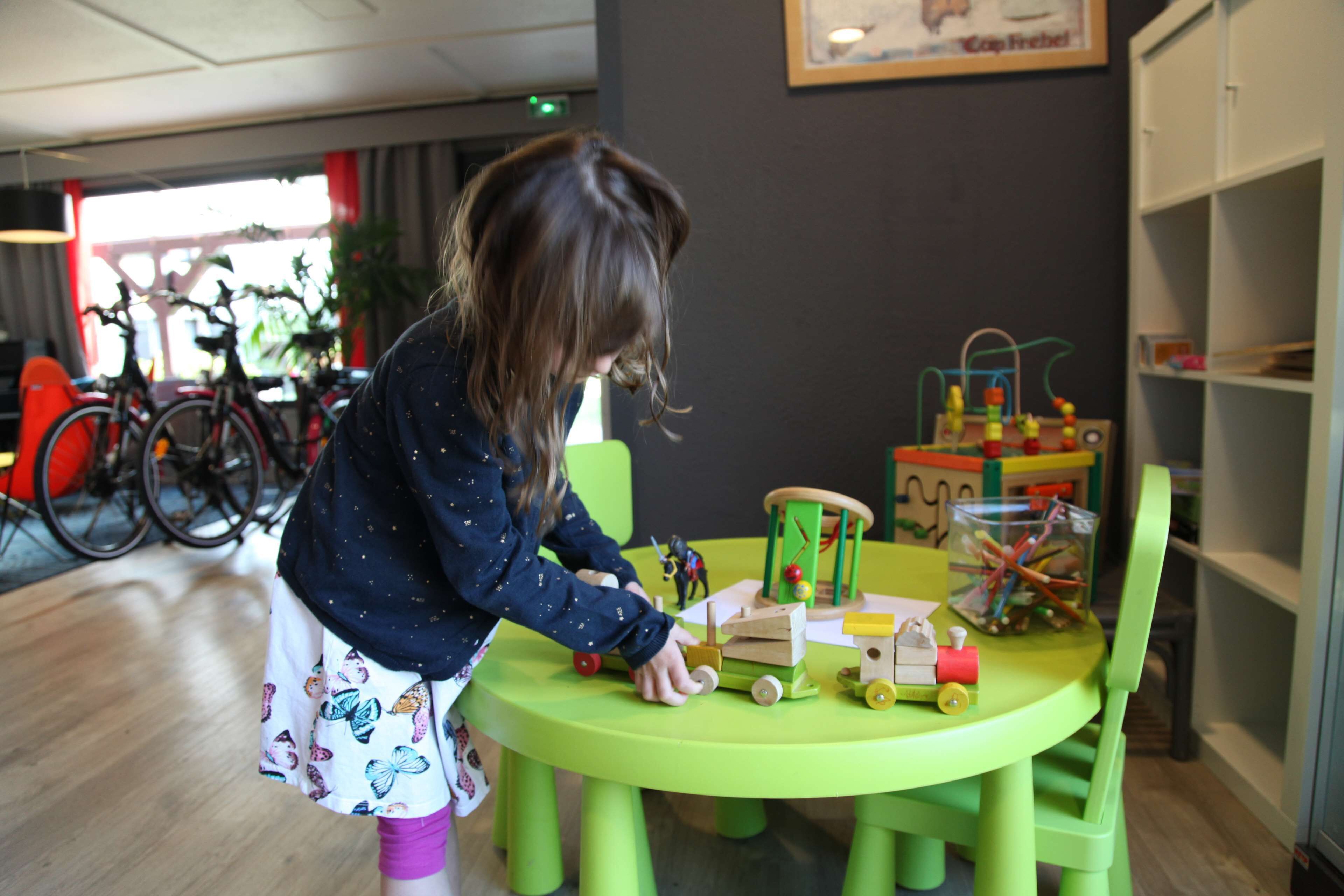 Un enfant joue avec des jouets sur une table verte dans une pièce où l’on aperçoit des vélos à l’arrière-plan.