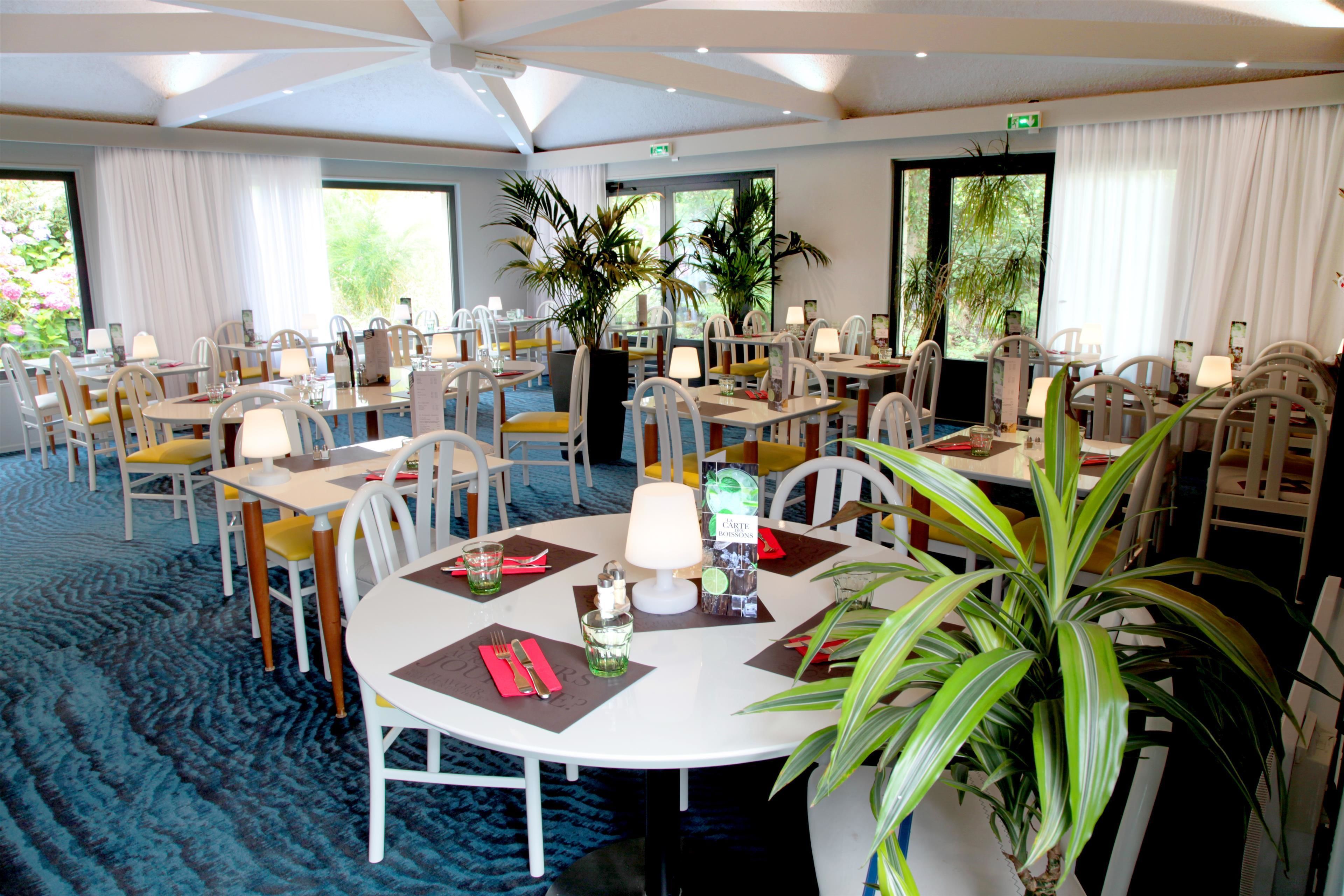Ein Hotelrestaurant mit runden Tischen, weißen Stühlen und Topfpflanzen.