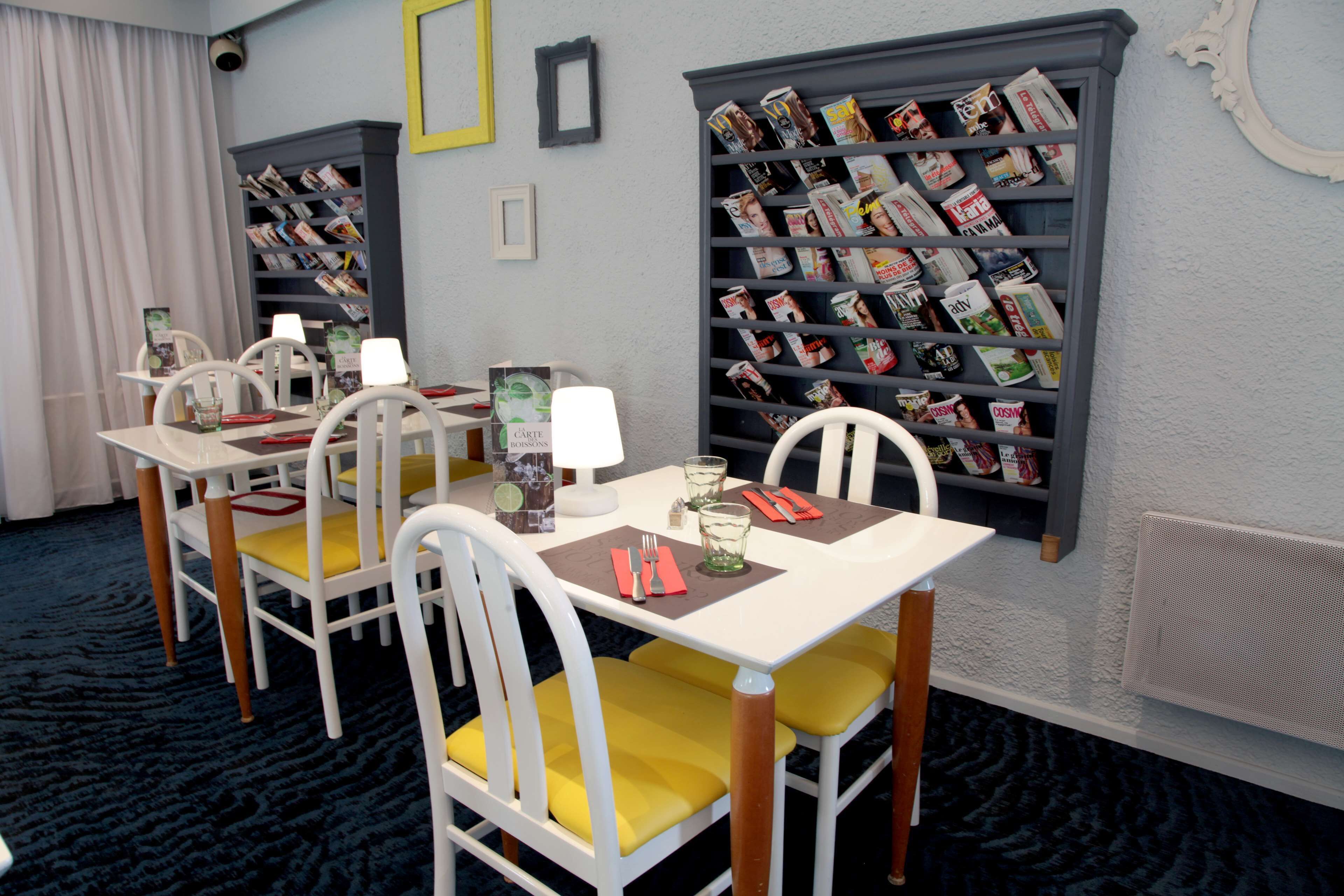 Un comedor con mesas y sillas blancas, una estantería llena de revistas y una ventana con cortinas.