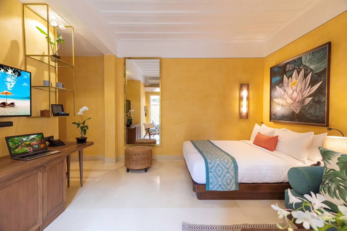 A hotel room with a bed, a desk with a laptop, a TV, and a large flower painting on the wall.