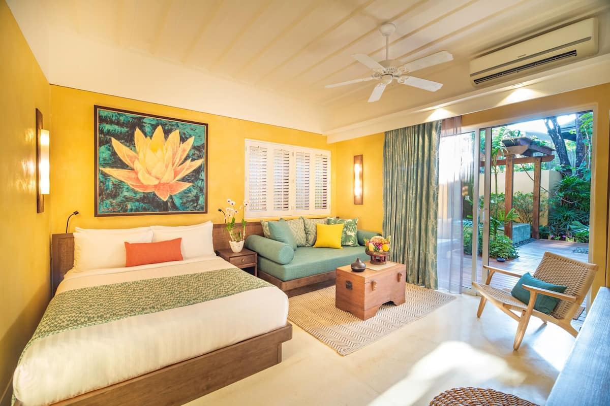 A hotel room with a bed, a sofa, a wooden chest, a chair, and a view of the garden through the glass door.
