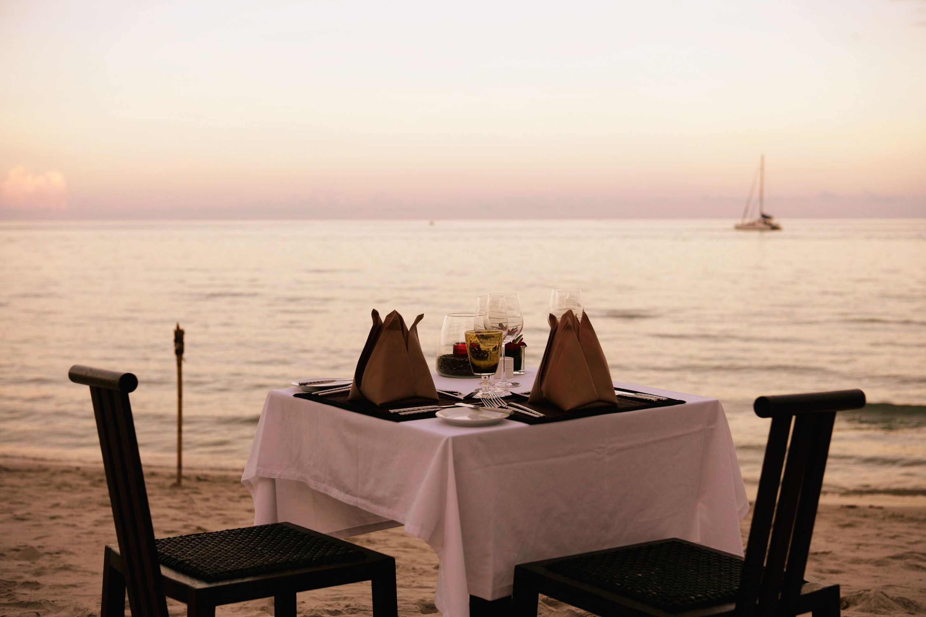 A table set for two on a beach with a view of the ocean and a sailboat in the distance.