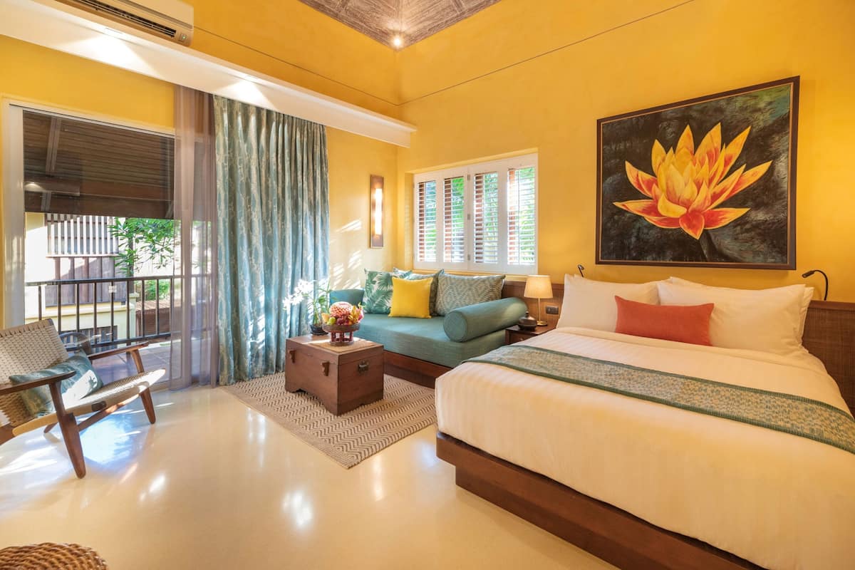 A hotel room with a bed, a sofa, a chair, a small table with a fruit bowl, and a painting of a lotus flower.