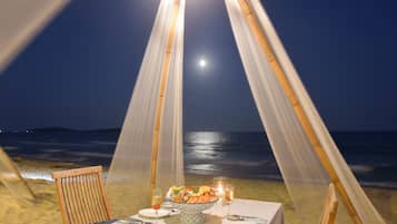 A beachside dining setup with a table, chairs, and a bowl of food, under a canopy with a view of the ocean and a moon in the sky.