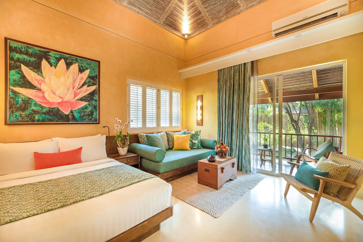 A hotel room with a bed, a sofa, a wooden chest, and a chair. There is a window with curtains and a view of greenery outside.