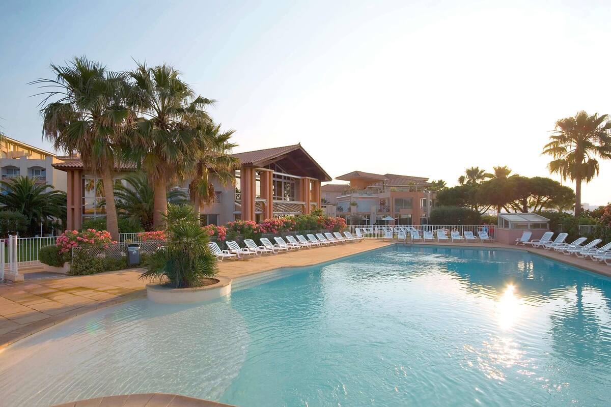 A swimming pool with lounge chairs, palm trees, and a building with balconies in the background.