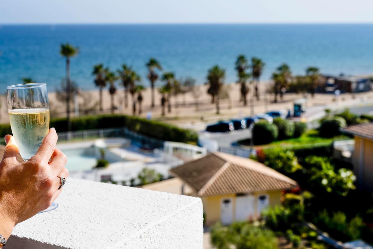 A hand holding a glass of white wine with a view of a beach and ocean in the background.