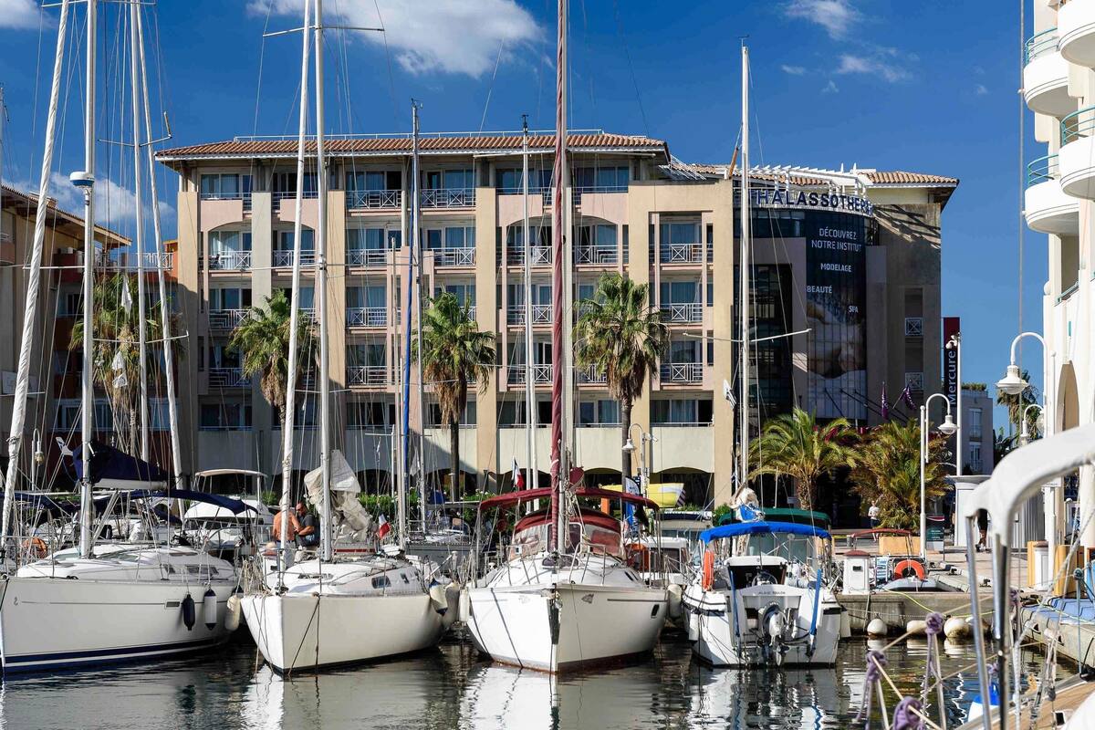 A marina with multiple sailboats docked, a building with balconies, and palm trees.