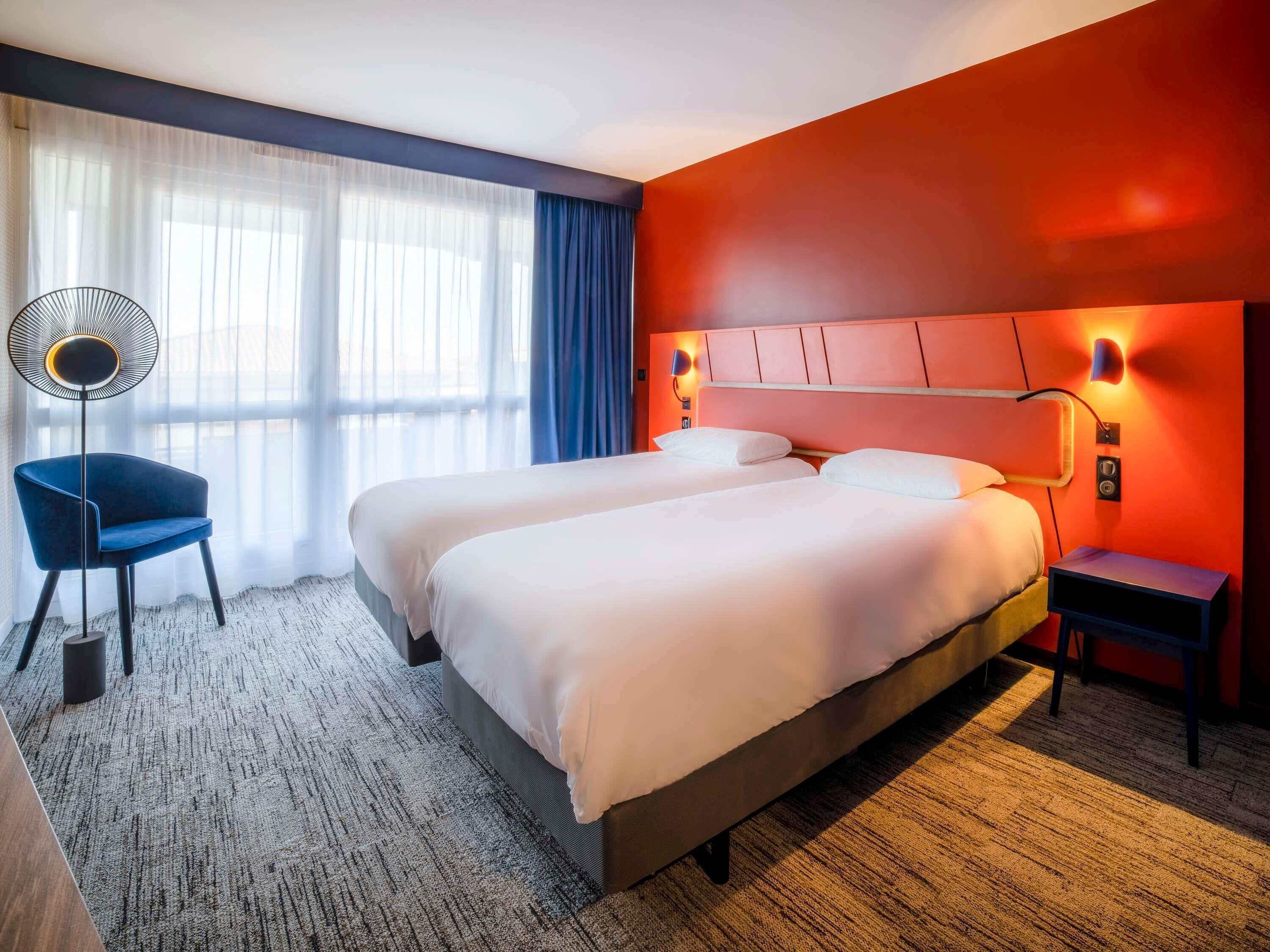 Ein Hotelzimmer mit zwei Betten, einem Sessel, einem Schreibtisch, einer Lampe und einem Fenster mit Vorhängen.