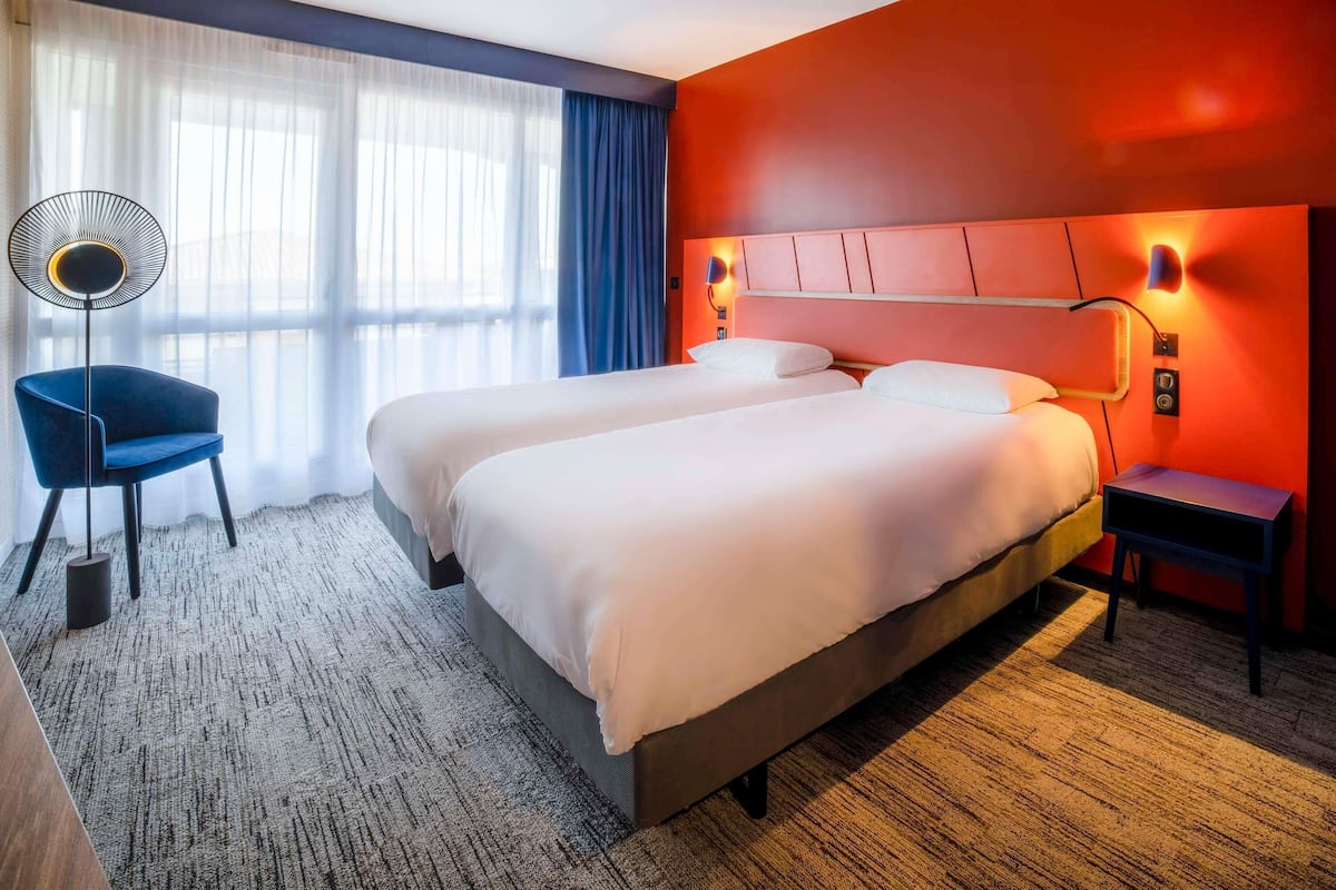 Ein Hotelzimmer mit zwei Betten, einem Sessel, einem Schreibtisch, einer Lampe und einem Fenster mit Vorhängen.