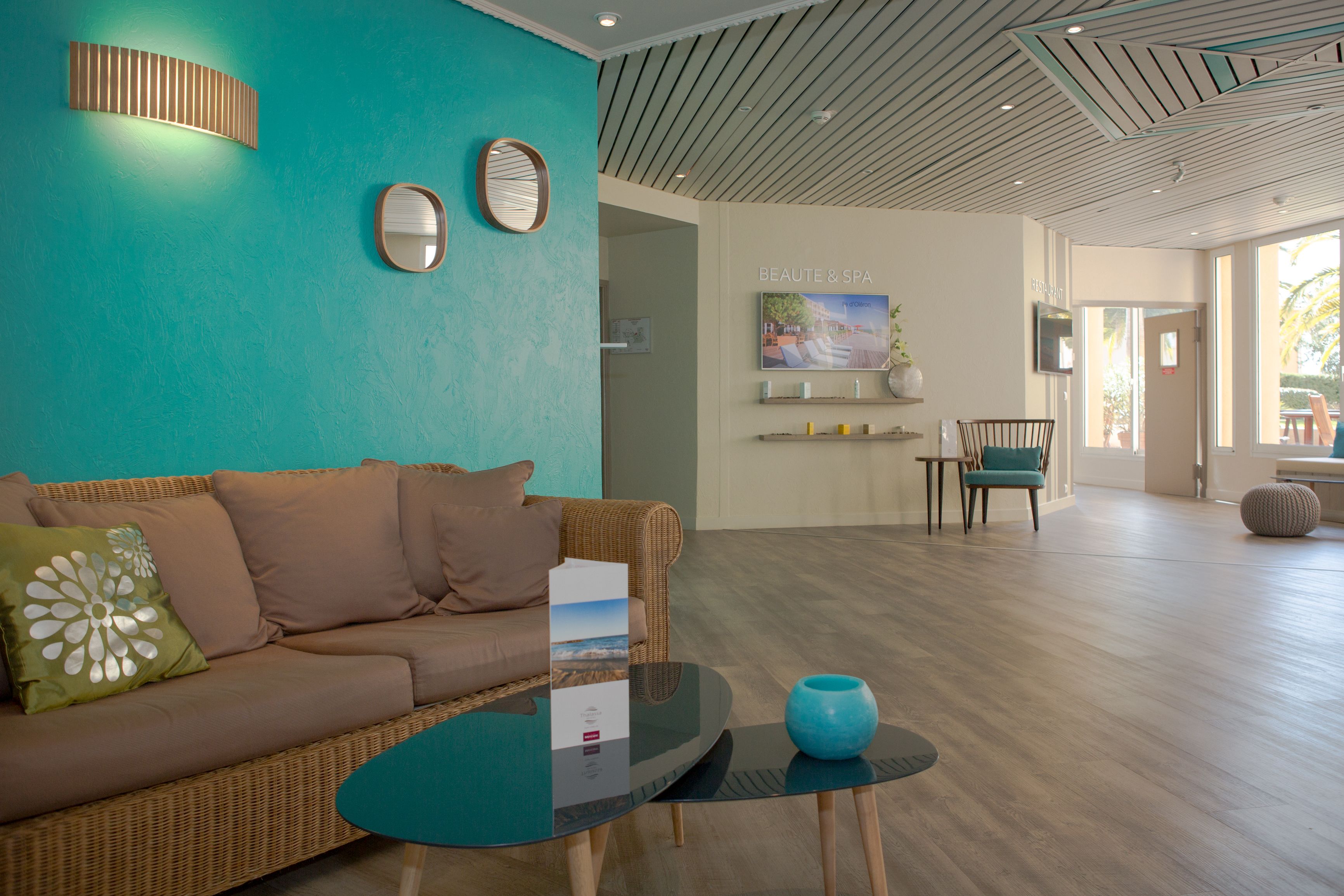 A modern hotel lobby with a teal wall, a wicker sofa, a glass coffee table, and a TV screen displaying 'BEAUTÉ & SPA'.