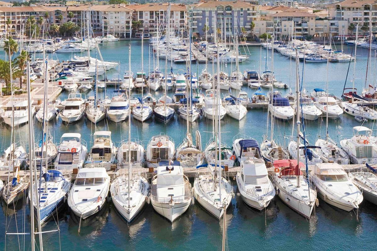 A marina with numerous boats docked, surrounded by buildings with balconies and a mountainous backdrop.
