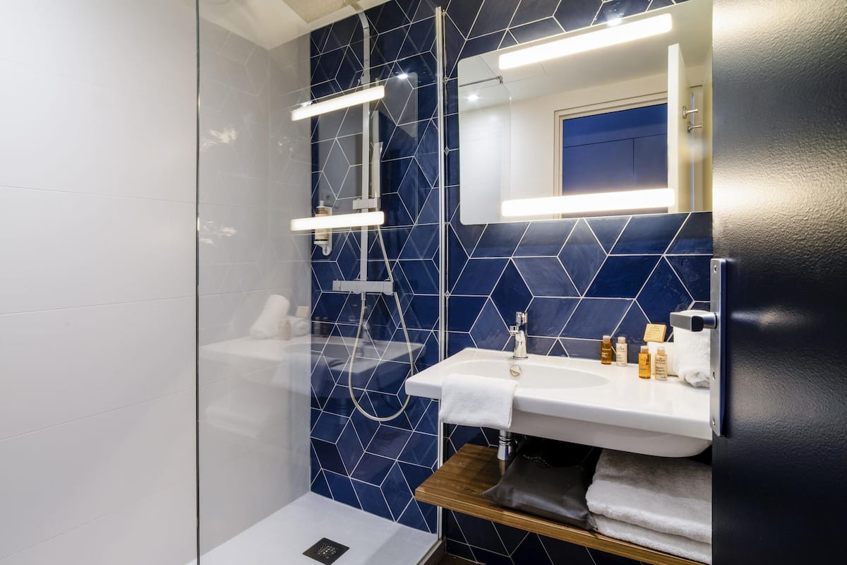 A modern bathroom with a glass shower enclosure, a white sink with a wooden shelf, and a blue-tiled wall.