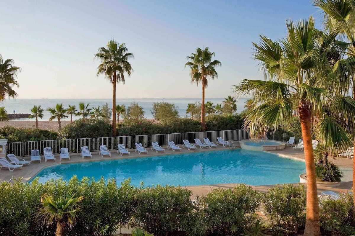 A pool with lounge chairs, palm trees, and a view of the sea.