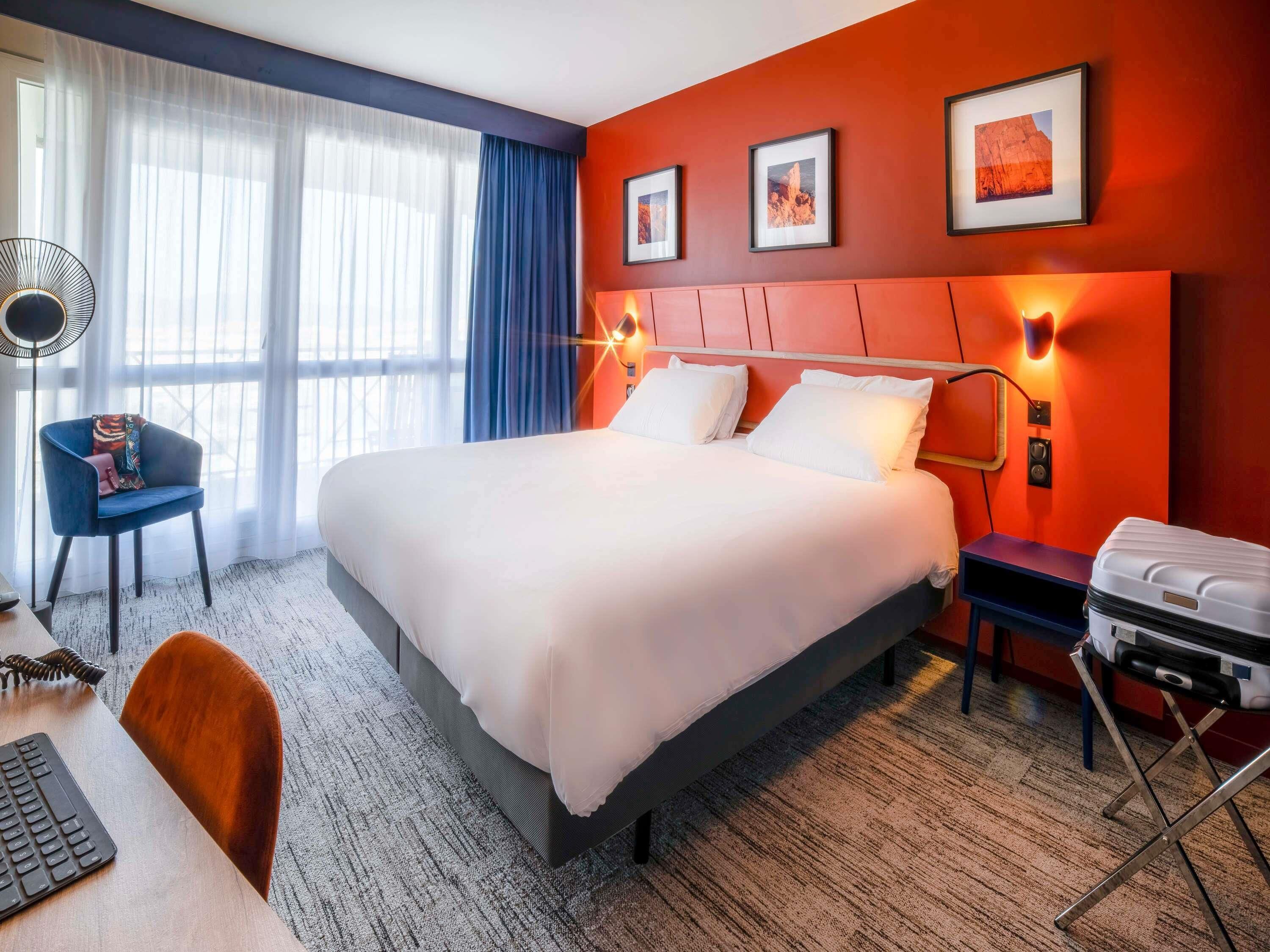 Ein Hotelzimmer mit einem großen Bett, einem Schreibtisch mit Tastatur, einem Sessel, einem Koffer und gerahmten Bildern an der Wand.